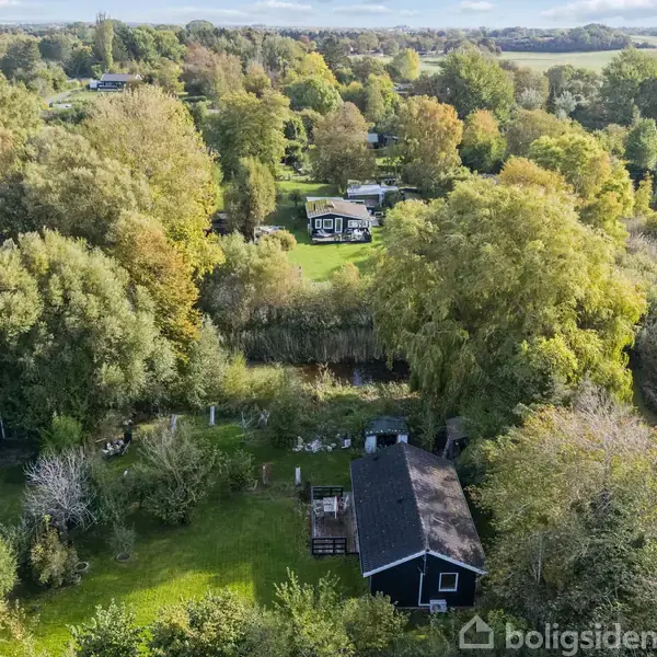 Et sort hus ligger blandt træer og buske i en frodig, grøn landskab. Omkring er der flere små bygninger, og i baggrunden ses marker og skovområder under en blå himmel.