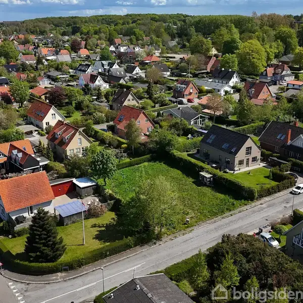 Huse med røde tage står stille i en villakvarter omgivet af grønt græs og træer under en klar blå himmel.