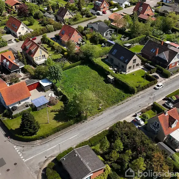 Huse med røde tage står side om side langs en vej i et beboelseskvarter. Omkring dem er grønne haver og træer. Trafikken er rolig med enkelte parkerede biler.