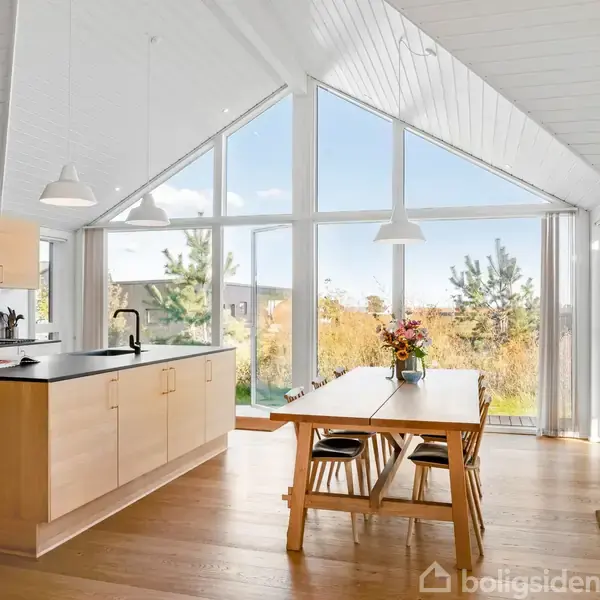 Køkken med træskabe står mod en stor glasvæg, der giver udsigt til en have. Et spisebord med blomster er placeret mellem køkkenøen og vinduerne.