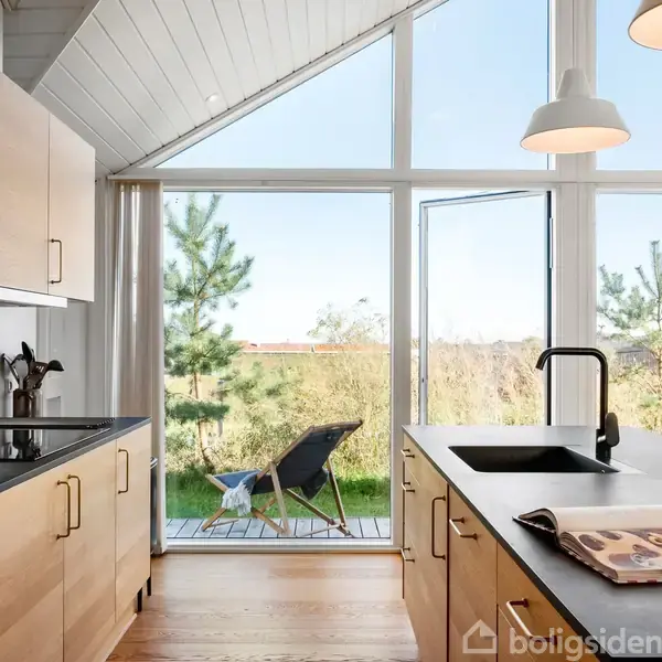 Et moderne køkken med træskabe og en sort bordplade; store vinduer åbner mod en terrasse med en stol. Udsigt til natur med træer og buske udenfor.