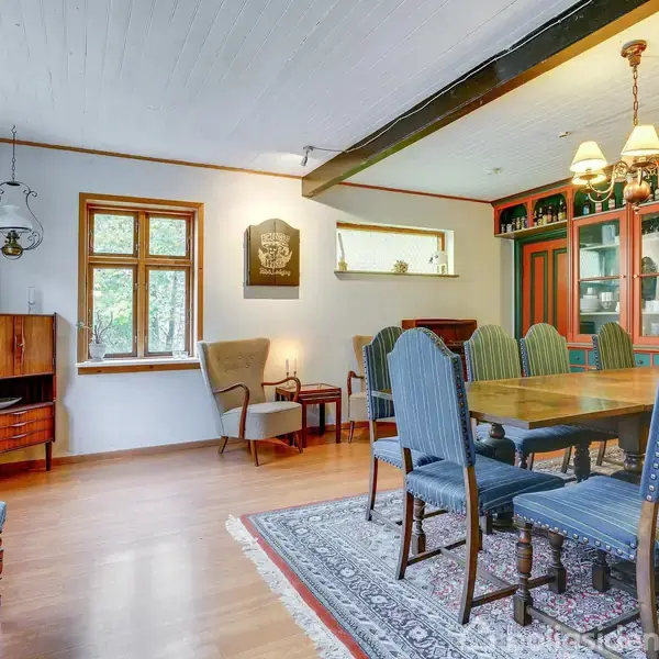 Spisestue med et langbord og syv stole omkring det. En lysekrone hænger over bordet. Baggrunden har skabe med glaslåger, mens der ses vinduer til venstre.