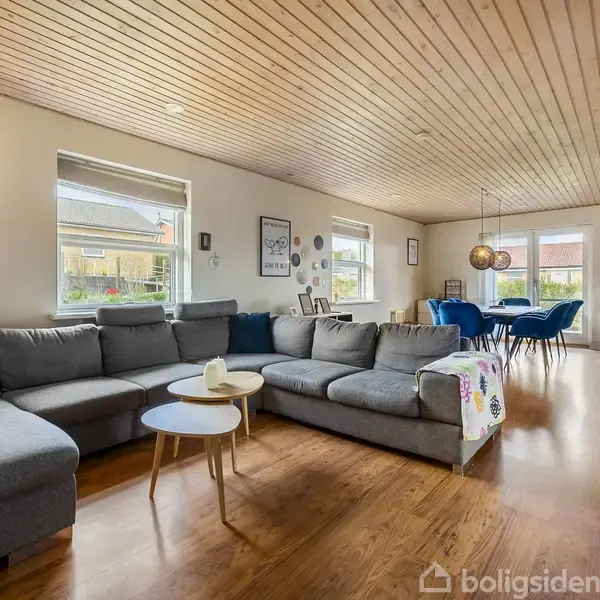Gråt sofaarrangement står stille i en stor, lys stue med trægulv og træloft. Et spisebord med blå stole står ved vinduer. Hyggelig og moderne atmosfære.