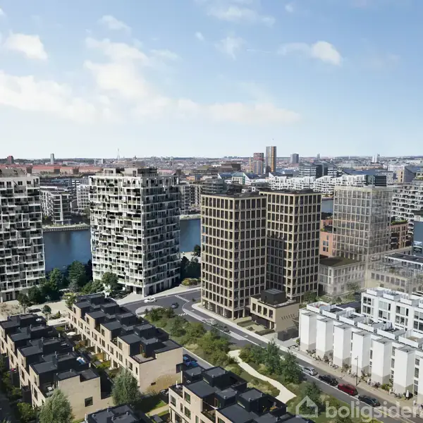 Flere høje bygninger står langs en flod i et byområde, omgivet af lavere bygninger og grønne områder under en klar blå himmel.