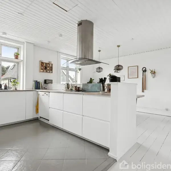 Et moderne køkken med hvide skabe og en central emhætte. Køkkenet har store vinduer, der giver naturligt lys, og rummet er indrettet med planter og køkkenudstyr.