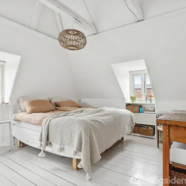 En seng står midt i et lyst, hvidmalet loftværelse med skrå vægge, omgivet af vinduer, planter og et træbord til højre.