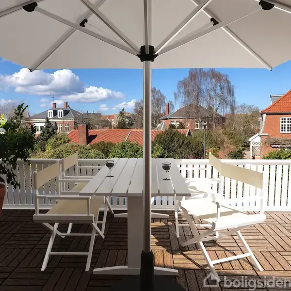 En udendørs terrasse med et hvidt bord og fire stole under en stor parasol. Et glas vin står på bordet. Omkring er der en hvid træhegn og grønne planter.
