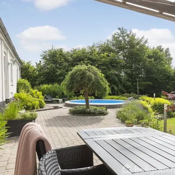 Terrassemøbler står på en flisebelagt terrasse, omgivet af grønne planter. I baggrunden ses en swimmingpool og frodige træer, der skaber en fredelig haveatmosfære under en blå himmel.