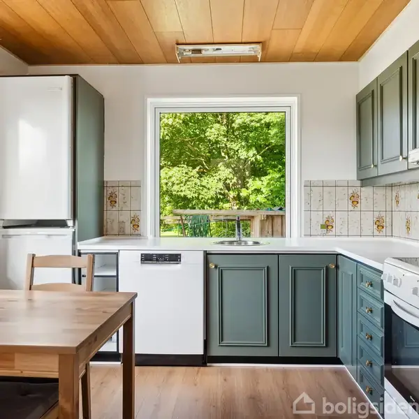 Køkken med lysegrønt skab og hvidt køleskab stående ved et vindue. Træbord med stole foran. Vindue viser udsigt til frodigt grønne træer udenfor.