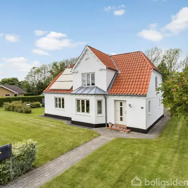 Hvidt hus med rødt tag står på en velplejet græsplæne med en gangsti og et træ ved siden. Blå himmel og få skyer i baggrunden.