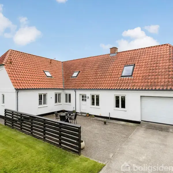 Hvidt hus med rødt tegltag står stille ved flisebelagt gårdsplads; lille terrasse med bord og stole. Græsareal omgiver gårdspladsen, og himlen er blå med spredte skyer.