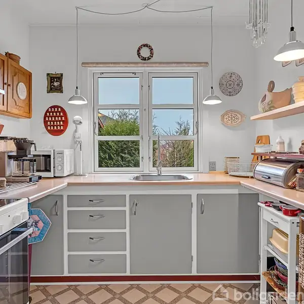 Et køkken med en grå-hvidvask, der udfører madlavningsfunktioner. Omgivet af hvide skabe, hængende lamper, små køkkenredskaber og udsigt til en grøn have gennem vinduet.