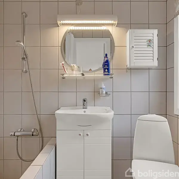 Vaskeskab med spejlrefleksion over og hylder med toiletartikler. En bruser er til venstre ved siden af badekarret. Toilet th. Fliseklædte vægge og et vindue lader lys ind i rummet.