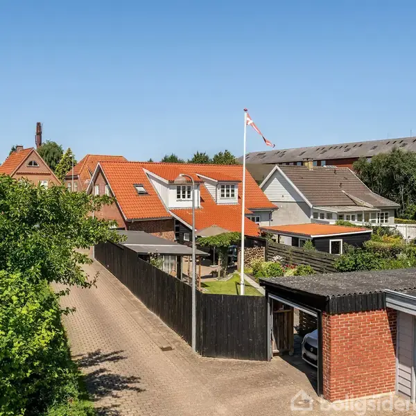 Flere huse med røde tage står langs en vej. Et flag blafrer i vinden i forgrunden, omgivet af grønne træer og buske.