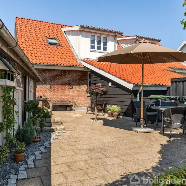 Terrasse med flisebelægning, hvor et bord med stole og en parasol står. Omgivet af planter og en murstensbygning med rødt tegltag i en solrig gårdhave.