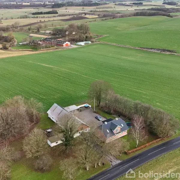 Et landsted med flere bygninger omringes af træer, beliggende på en stor, grøn mark. En vej løber tæt forbi ejendommen. I baggrunden ses flere åbne landskaber og småbygninger.