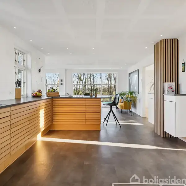 Et moderne køkken med en træbeklædt køkkenø udstyret med barstole, placeret i et lyst rum med store vinduer og en udsigt til en have eller skov.