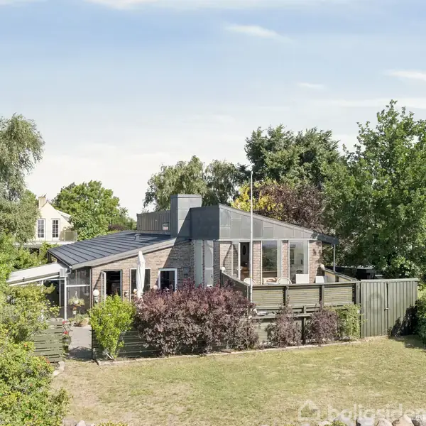 Et moderne hus med store vinduer står solbeskinnet i en grøn have. Omgivelserne inkluderer træer og buske, hvilket skaber en hyggelig og naturlig atmosfære.