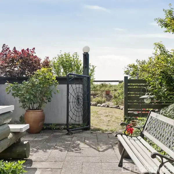 Havebænk står ved siden af et træbord på en terrasse med fliser. Bagved er et hegn med en dekoreret metaldør omgivet af forskellige buske og træer.