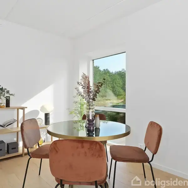 Et rundt bord med blomsterarrangement omgivet af fire brune stole står i et lyst rum med stort vindue ud til en have. Reol til venstre med bøger og planter.