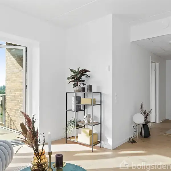 Hylde med planter og dekorationer står mod en hvid væg, mens en åben dør fører til en balkon med udsigt til åbent landskab. En blå sofa står i nærheden.