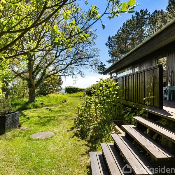 Sommerhus i træ ligger på en grøn græsplæne omgivet af høje træer og buske. Der er en terrasse med borde og stole foran huset. Skovklædt landskab i baggrunden.