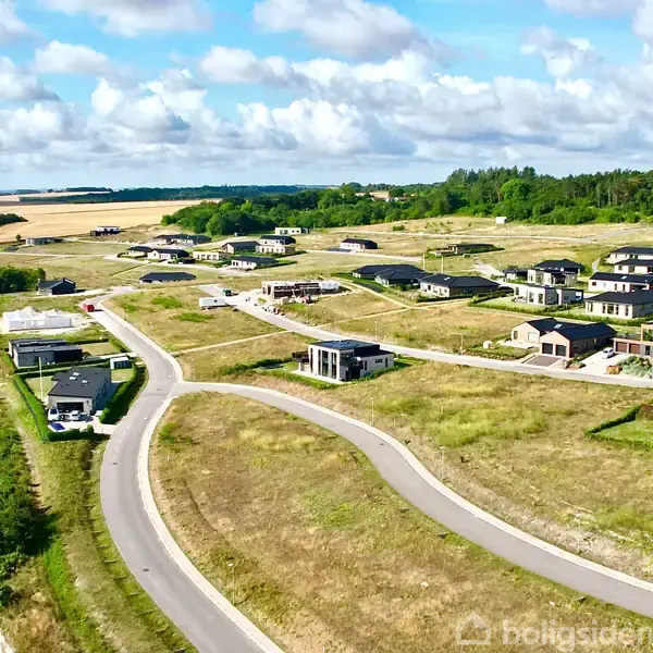 Flere moderne huse spredt langs snoede veje i et nybygget boligområde omgivet af grønne marker og skove under en delvist skyet himmel.