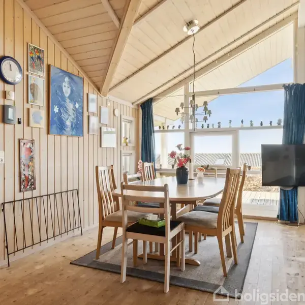 Spisebord med seks stole står i et træbeklædt rum med store vinduer, der lader naturligt lys strømme ind. Væggene er dekoreret med billeder og en urtavle.