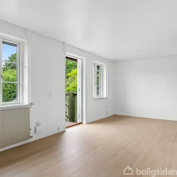 Et lyst rum med hvide vægge og trægulv har vinduer og en glasdør til en balkon. Udenfor ses grønne træer. Rummet er tomt og har en radiator.