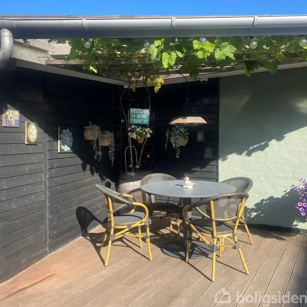 Terrasse med træmøbler og parasol på gule fliser. Omkranset af hvidmalet husvæg med dekorationer. Planter og buske langs kanten, og klar himmel i baggrunden.