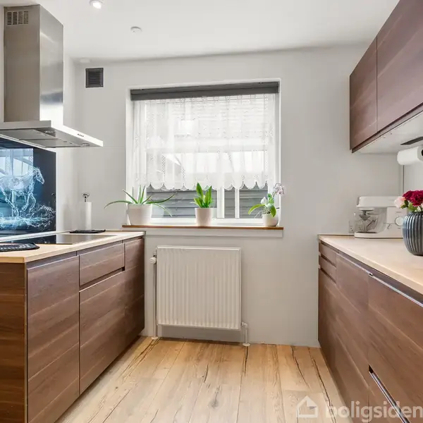 Et moderne køkken med træskabe og trægulv. Rummet er lyst med en central vindueskarm med planter. Der er en ovn og emhætte på venstre side.