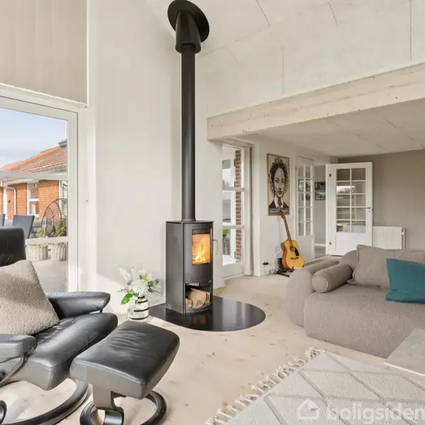 Brændeovn opvarmer en moderne stue med sorte lænestole og grå sofa; udendørs lys skinner ind gennem store vinduer, og en guitar står ved siden af et portræt.