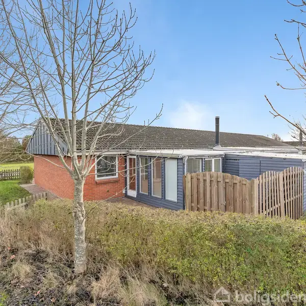 Et rødt murstenshus med blå træbeklædning står omgivet af en grøn have med nøgne træer. En lille træhegn sektion afgrænser et område ved huset. Himmel med lette skyer.