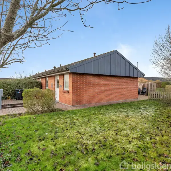 Et rødt murstenshus med sort tag står i en have omkranset af hække og træer. En sti fører op til huset, omgivet af græsplæne og buske.