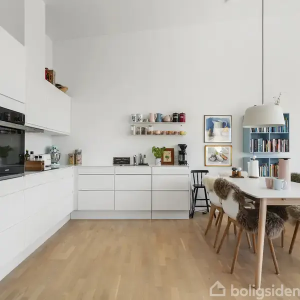 Køkken med hvide skabe og integreret ovn, står i et moderne rum. Ved siden af er der et spisebord med stole, dekoreret med en pelset pude, under en hængelampe.