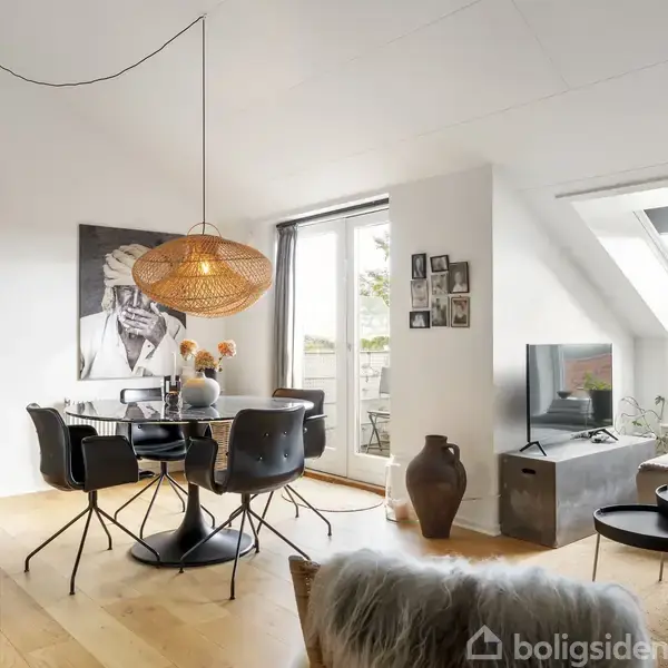 En moderne stue med spisebord og sorte stole, hvor en stor loftlampe hænger over bordet. Billeder og planter dekorere væggene, mens et fjernsyn står ved et skråvindue.