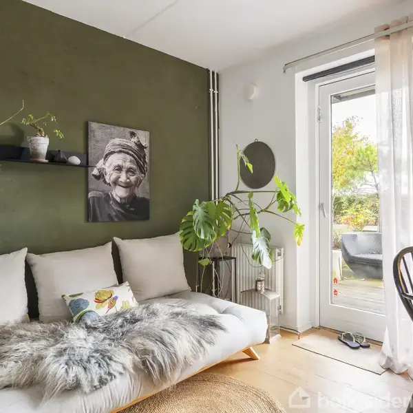 Sofa med hvide puder og et pelstæppe står mod en grøn væg med billedkunst. Ved siden af er en plante og en glaspendel. En dør åbner til terrasseområdet.