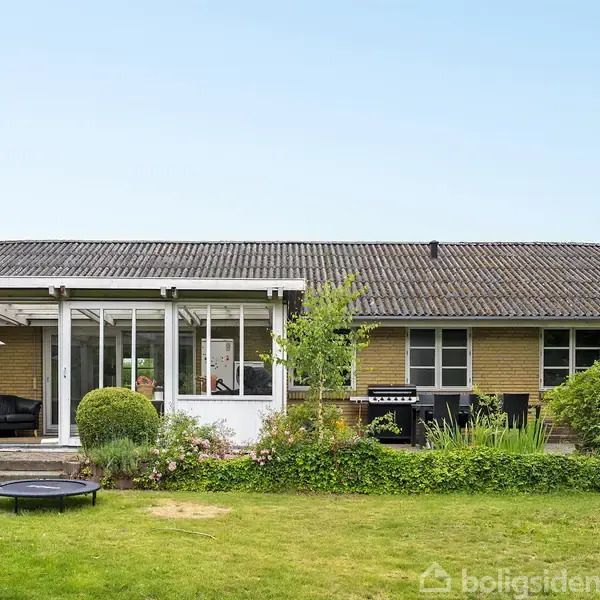 En enkeltstående étplans gule murstensvilla står i en grøn have med en græsplæne. Huset har en glasoverdækket terrasse og en grill ved siden af farverige buske.