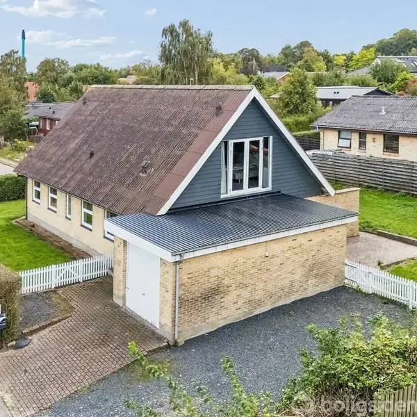 Et parcelhus med en stor tagkonstruktion og en tilknyttet garage. Huset er omgivet af en velplejet have med hække og grusindkørsel. Det ligger i et villakvarter med flere lignende huse.