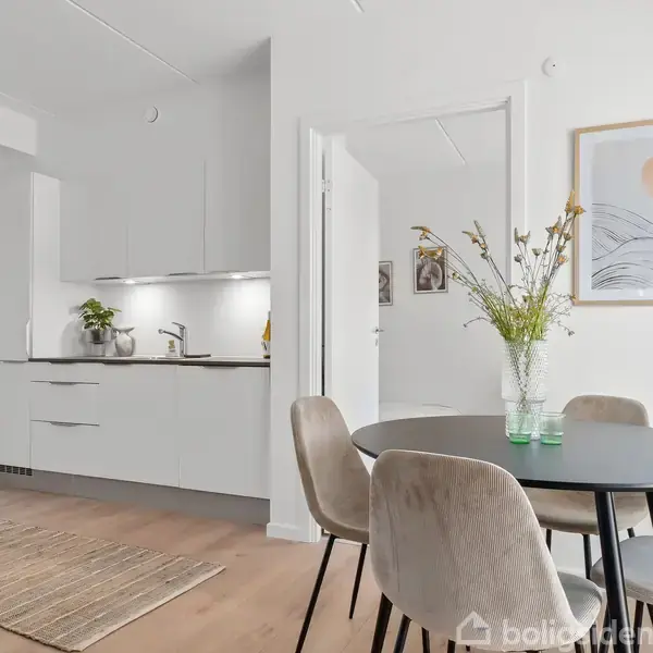 Et rundt spisebord med fire stole står i centrum med blomster i en vase. Køkkenet er moderne med hvide skabe. Lys træfarvet gulv og vægkunst skaber en minimalistisk atmosfære.