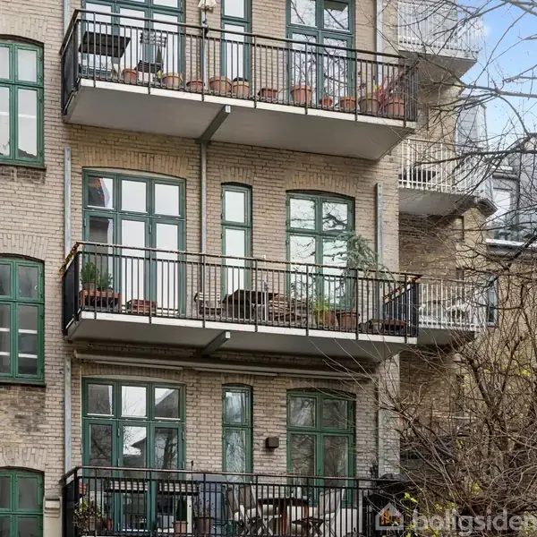 En murstensbygning med grønne vinduer og altaner, hvor blomsterkasser er placeret. Træer uden blade omkranser bygningen, hvilket antyder en vinter- eller efterårssæson.