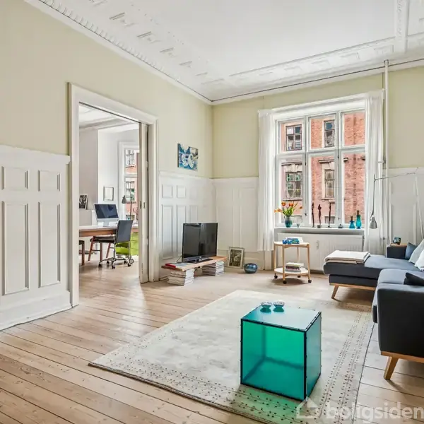 Stue med gråt sofaarrangement står på et tæppe, omgivet af lyse trægulve og cremevægge. En stor vindue lader naturligt lys ind, og åbningen fører til et arbejdsområde.