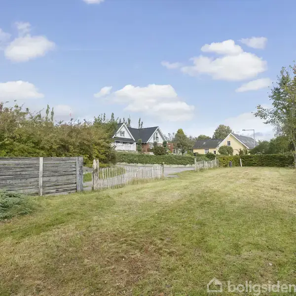 Græsplæne strækker sig ned ad en blød skråning, flankeret af træer og buske. I baggrunden ses to huse med stejle tage under en klar himmel med spredte skyer.