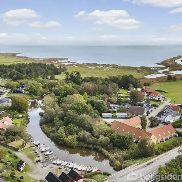 Små både ligger fortøjet langs en slyngende kanal, omgivet af huse med røde tage og grønne træer; i baggrunden ses åbne marker og havet under en let skyet himmel.