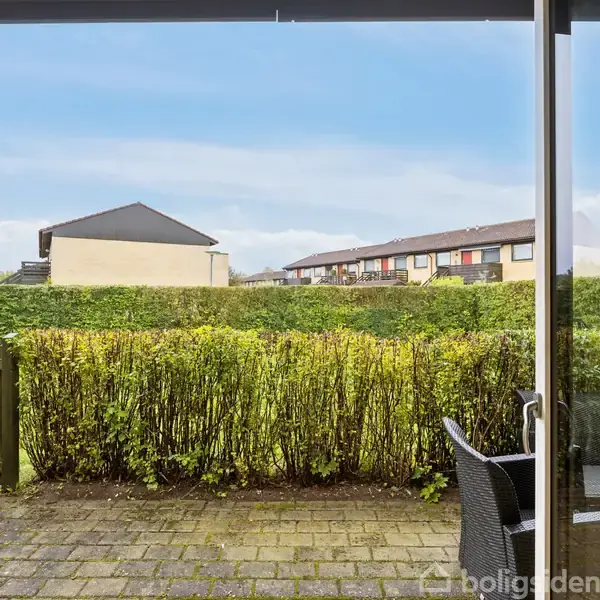 Terrassemøbler står på en flisebelagt terrasse foran en høj hæk. I baggrunden ses rækkehuse og en blå himmel med lette skyer. En havelåge forbinder terrassen med haven.