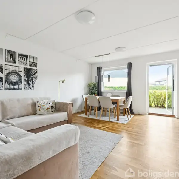 Sofa i en stue vender mod et TV; spisestue med bord og stole ved fransk dør. Vægkunst af Eiffeltårnet til venstre, lys trægulv.
