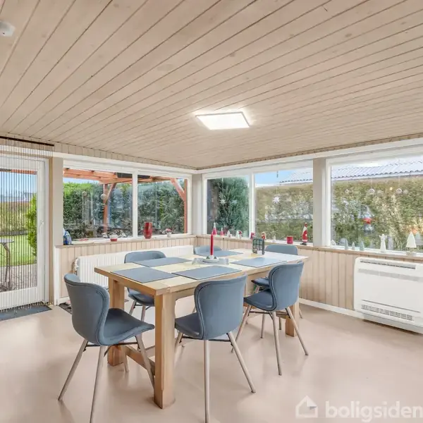 Et lyst spisestueområde med et træbord omgivet af blå stole. Der er store vinduer, som giver udsigt til en have. Rummet har et lyst trægulv og paneleret loft.