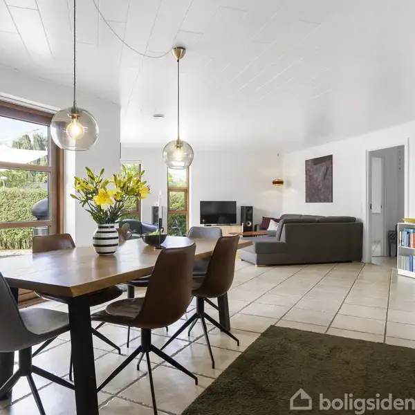 Et spisebord med blomster står i et lyst boligområde. Rummet har flisegulv, en grå sofa, mange vinduer, bøger på en reol og moderne belysning.