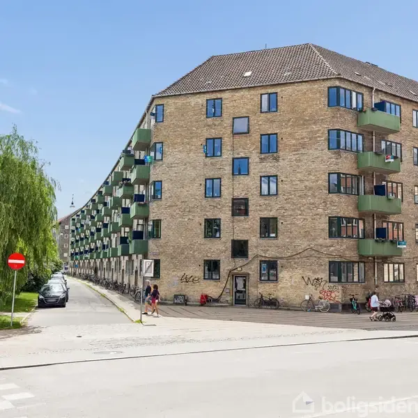 Et stort murstenslejlighedskompleks med flere etager står på hjørnet af en gade. Bygningen har grønne altaner. Foran bygningen er der parkerede cykler og et lille træ på fortovet.