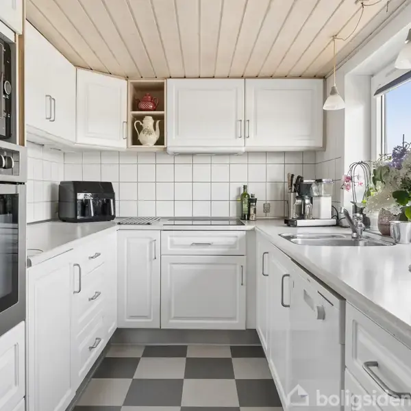 Hvidt køkken med fliser og træloft. Køkkenet indeholder en ovn, mikroovn, airfryer og blomster i vindueskarmen. Der er skakternet gulv, og skabene har moderne håndtag.
